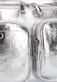 Zilveren metallic ritszak met een gladde textuur, duale compartimenten en glanzend zilver beslag. Beschikt over zichtbare stiksels en accenten.