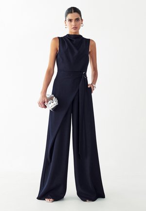 Kobieta ubrana w bezrękawnikowy czarny kombinezon z szerokimi nogawkami, trzymająca małą metaliczną kopertówkę, stojąca na tle jednolitego białego tła.