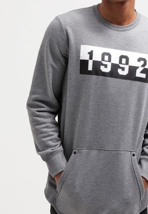 Grijze sweatshirt met een contrasterende zwart-witte grafiek met "1992." Heeft een ronde halslijn, lange mouwen en een voorkant zak.