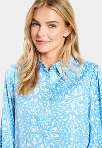 Kék gombos ing, amelyen fehér, bonyolult virág- és paisley minta található, hosszú ujjakkal és laza fitten. Sima textúra.