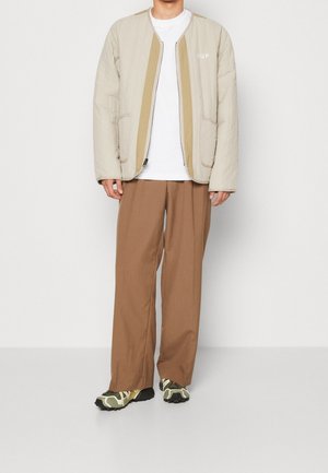 Beżowa pikowana kurtka z beżowymi akcentami, biała koszulka, brązowe szerokie spodnie i zielone sneakersy w wzory. Prosty design z kieszeniami.