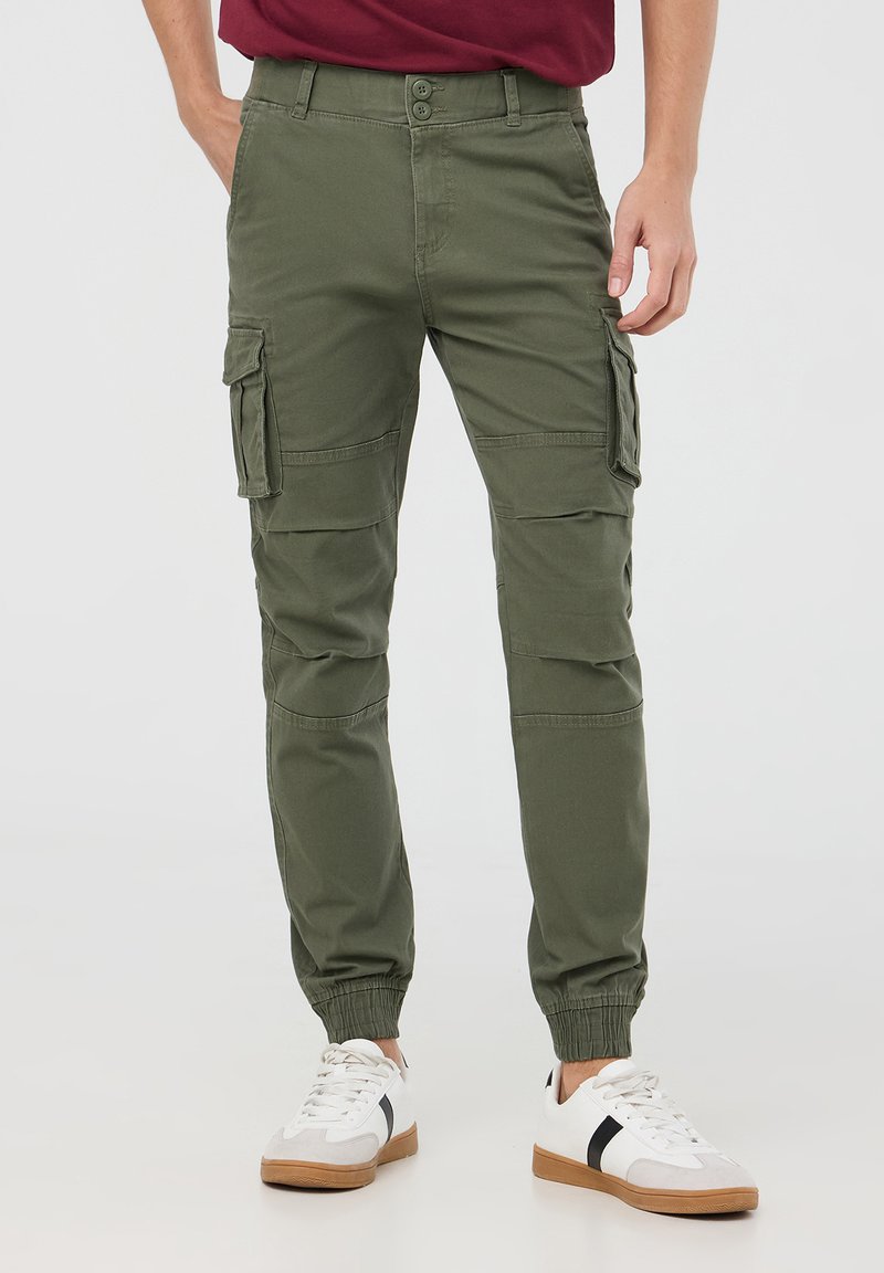 Uomo in pantaloni cargo verdi con polsini elasticizzati e sneakers bianche con strisce nere, in piedi contro uno sfondo neutro.