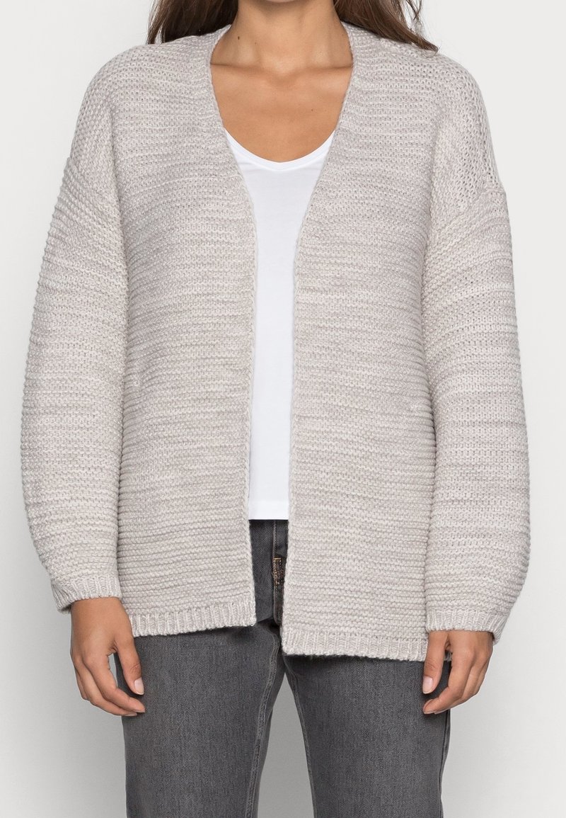 Jasnoszara, dziergana kardiganka o luźnym kroju, z otwartym przodem i szerokimi rękawami. Teksturowane, prążkowane wykończenia u dołu i na mankietach; noszona na białą bluzkę i dżinsy.