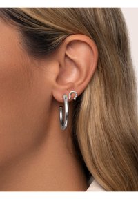 Pendientes de aros plateados y pequeños pendientes en forma de herradura, con un acabado brillante. El aro es grande, mientras que el pendiente de acento tiene piedras incrustadas.