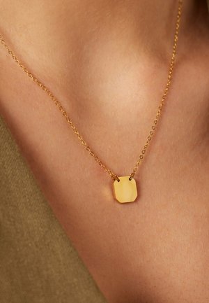 Collier en chaîne d'or avec un petit pendentif octogonal lisse reposant sur la peau nue près de la clavicule.