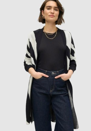 Cardigan a righe bianche e nere con maniche lunghe, design aperto e texture a maglia, indossato sopra una maglietta nera e jeans a vita alta blu.