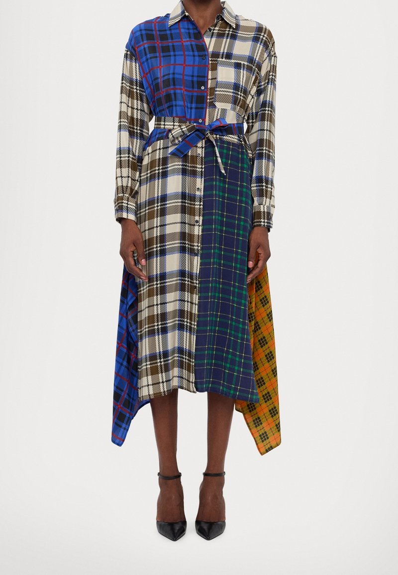 Femme portant une robe patchwork à manches longues avec ceinture, à carreaux avec des motifs bleus, verts, marron, blanc et orange, et des talons noirs à bride à la cheville.