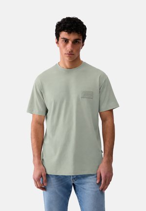 Uomo con capelli corti e ricci che indossa una maglietta verde chiaro a girocollo e jeans blu, in piedi contro uno sfondo bianco neutro.