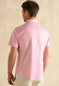 Ružová košeľa s krátkym rukávom a špicatým golierom, hladká na dotyk, s detailom na štítku na zadnej strane; kombinovaná so svetlými nohavicami.