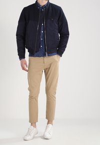 Navy bomber jacket with zip closure, paired with a blue patterned shirt and khaki chinos. White sneakers complete the outfit.