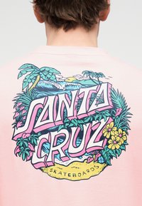 Πίσω μέρος ενός ανοιχτό ροζ πουκάμισου με πολύχρωμο γραφικό "Santa Cruz Skateboards" που απεικονίζει φοίνικες, κύματα του ωκεανού, λουλούδια και παραλιακή σκηνή.