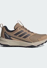 Trail running shoes featuring a tan and black design, textured upper, sturdy rubber outsole, and distinctive three-stripe branding.