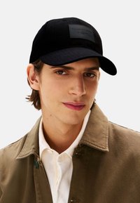 Gorra de béisbol negra con un acabado texturizado y una placa rectangular de cuero en la parte frontal. Se lleva con una camisa clara y una chaqueta marrón.