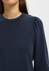 Top a maniche lunghe blu navy con scollatura rotonda e dettagli arricciati sulle spalle, realizzato in un tessuto leggero e liscio.