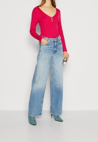 Frau trägt ein leuchtend pinkes Langarmoberteil mit Knöpfen, weit geschnittene hellblaue Jeans, türkisfarbene Absatzstiefel und hält eine metallische Clutch.