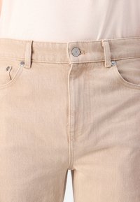 Jeans de mezclilla beige con un diseño clásico de cinco bolsillos, cierre de botón metálico y sutiles detalles de costura. Tejido suave y texturizado.