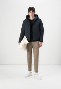 Schwarze gefütterte Kapuzenjacke, beige Hose, weiße Sneakers. Das Model hält eine cremefarbene Tasche und steht vor einem schlichten hellen Hintergrund.