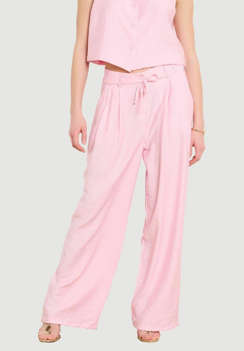 Person trägt lockere, hoch taillierte, hellrosa Hosen mit gebundenem Bund, kombiniert mit passendem cropped Top und Sandalen mit Absatz.