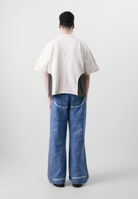 Beige Kurzarmoberteil mit grauem Rücken, kombiniert mit lockeren blauen Jeans, die verstreute weiße Sprenkel und ausgefranste Akzente aufweisen.