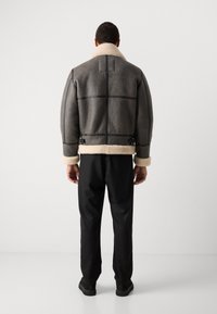 Γκρίζα βομβιστική ζακέτα με επένδυση από γούνα, καπέλο, μαύρες λεπτομέρειες και ρυθμιζόμενους φουσκωτούς ιμάντες στη μέση, συνδυασμένη με μαύρα παν trousers και παπούτσια.