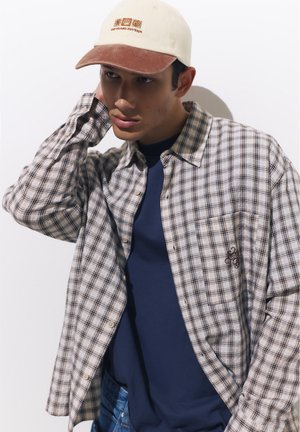 Young man wearing beige cap and plaid shirt over a navy blue t-shirt, posing with hand behind his neck against a plain white background.