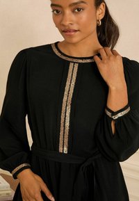 Schwarzes Kleid mit langen Ärmeln, das goldene Paillettenakzente entlang des runden Halsausschnitts und im Zentrum aufweist. Weicher Stoff und taillierter Gürtel.