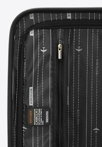 Intérieur d'une valise de voyage noire avec poche en maille, tissu à motifs avec des rayures blanches et des icônes d'avion, compartiments zippés et étiquette de marque.