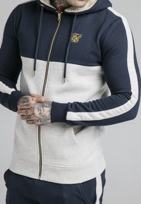 Felpa con zip di colore blu navy e grigio chiaro, con un logo dorato, dotata di cappuccio foderato, due tasche laterali e dettagli a righe sulle maniche.