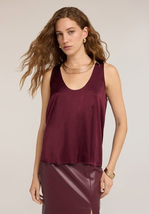 Vrouw met lang golvend haar draagt een mouwloze donkerrode satijnen top, bijpassende leren rok, gouden ketting, oorbellen en armbanden tegen een neutrale achtergrond.