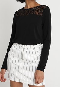 Svart långärmad topp med spetsdetaljer vid halsringningen, kombinerad med en vitrandig denimkjol med en framdragkedja och cirkulär accent.