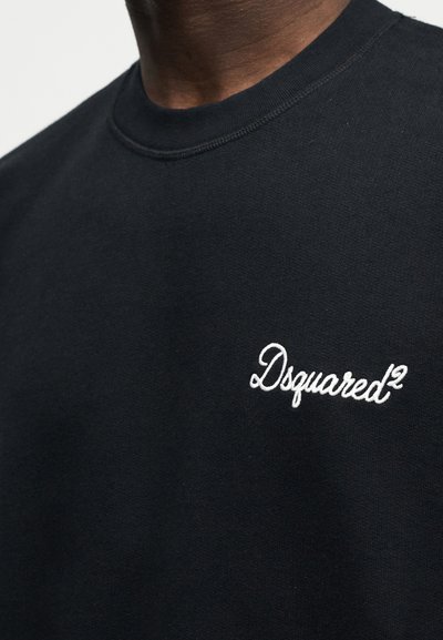 Κοντινό πλάνο ενός ατόμου που φοράε ι μαύρο μπλουζάκι με λευκό κεντημένο λογότυπο "Dsquared²" στο στήθος.