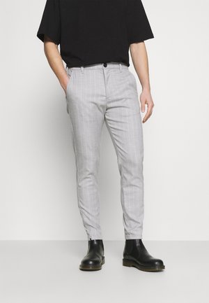 Mann trägt hellgraue Slim-Fit-Hose mit dezenten Streifen, ein schwarzes Hemd und schwarze Leder-Stiefeletten, steht mit einer Hand in der Tasche.