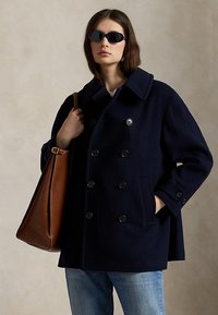 Frau trägt einen dunkelblauen zweireiher Mantel, hellblaue Jeans, dunkle Sonnenbrille und eine große braune Ledertasche auf der Schulter.