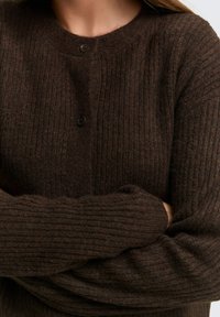 Brauner gerippter Strickpullover mit rundem Halsauschnitt und drei Knöpfen an der Vorderseite. Die Textur ist weich und dehnbar.