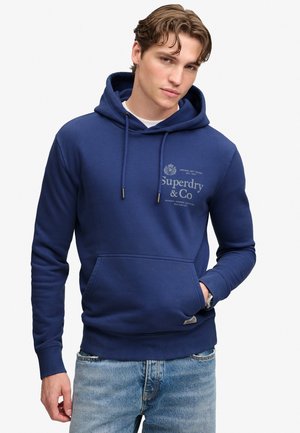 Młody mężczyzna ubrany w niebieską bluzę z kapturem Superdry z przednią kieszenią oraz jasnoniebieskie dżinsy, stoi z jedną ręką w kieszeni na białym tle.