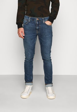 Mann trägt enge blaue Jeans mit umgeschlagenen Säumen, weiße Sneakers und ein dunkel getarntes Langarmhemd, steht vor einem schlichten Hintergrund.