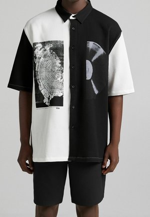 Mand iført sort-hvid kortærmet skjorte med knapper og tryk af træring og vinylplade, kombineret med sorte shorts.