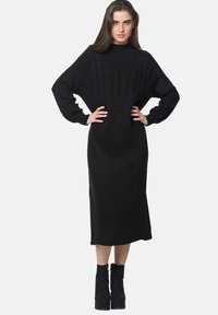 Robe noire tricotée avec un col haut, texture côtelée et manches longues. Elle a une silhouette ajustée, s'étendant jusqu'au mollet. Assortie avec des bottines noires.