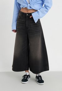 Calças de ganga largas pretas com um acabamento desgastado, combinadas com ténis pretos com detalhes brancos e uma camisa de botão às riscas azuis.