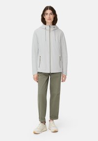 Vrouw staat rechtop, gekleed in een lichtgrijze gewatteerde zip-jas met capuchon, olijfgroene opgerolde broek en beige sneakers op een witte achtergrond.