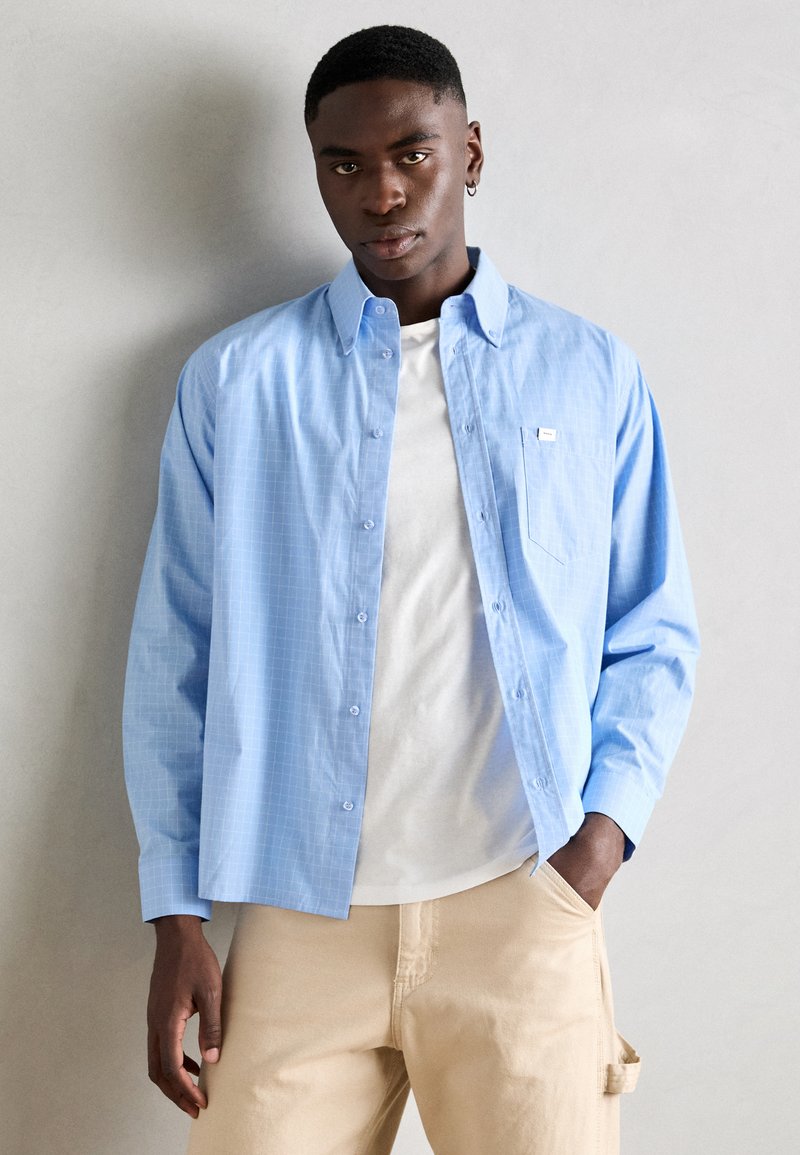 Lichtblauw geruit overhemd met lange mouwen en knopen, voorzien van een patch pocket. Draagt over een wit T-shirt en gecombineerd met beige broeken.