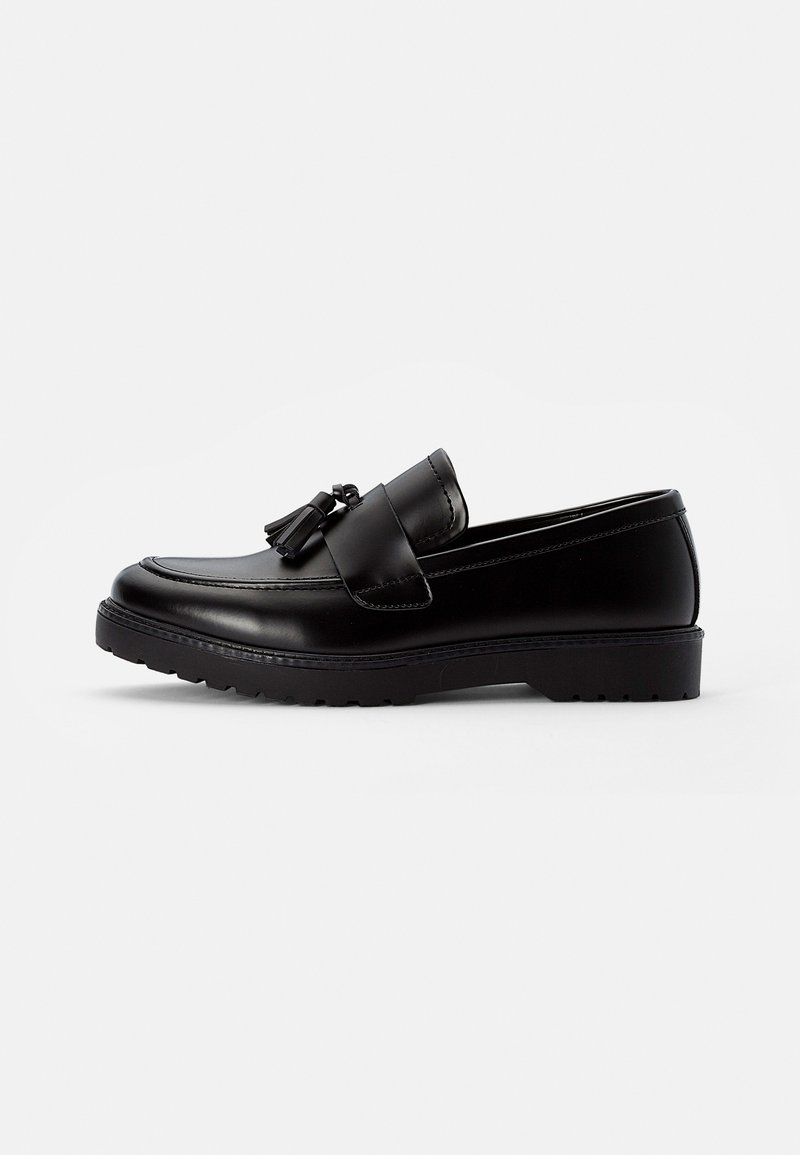 Mocassin à pampilles en cuir noir avec détails couture et semelle épaisse en caoutchouc sur fond blanc.