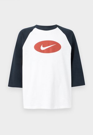 Hombre vistiendo una camiseta blanca y azul marino de tres cuartos de manga con un óvalo rojo y el logo blanco de Nike, jeans azul claro y una gorra de béisbol azul claro.