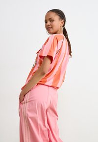 Camisa de manga corta a rayas en rosa y naranja con cuello redondeado, combinada con pantalones anchos rosas que tienen bolsillos laterales y tres rayas.