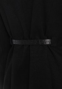 Svart klänning med omslag i en texturerad tyg, åtdragen vid midjan med ett smalt bälte i svart krokodilsmönster.