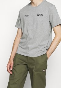 Homme portant un t-shirt Levi's gris avec un texte noir et un pantalon Dickies vert olive avec un patch logo sur la poche, les mains dans les poches.