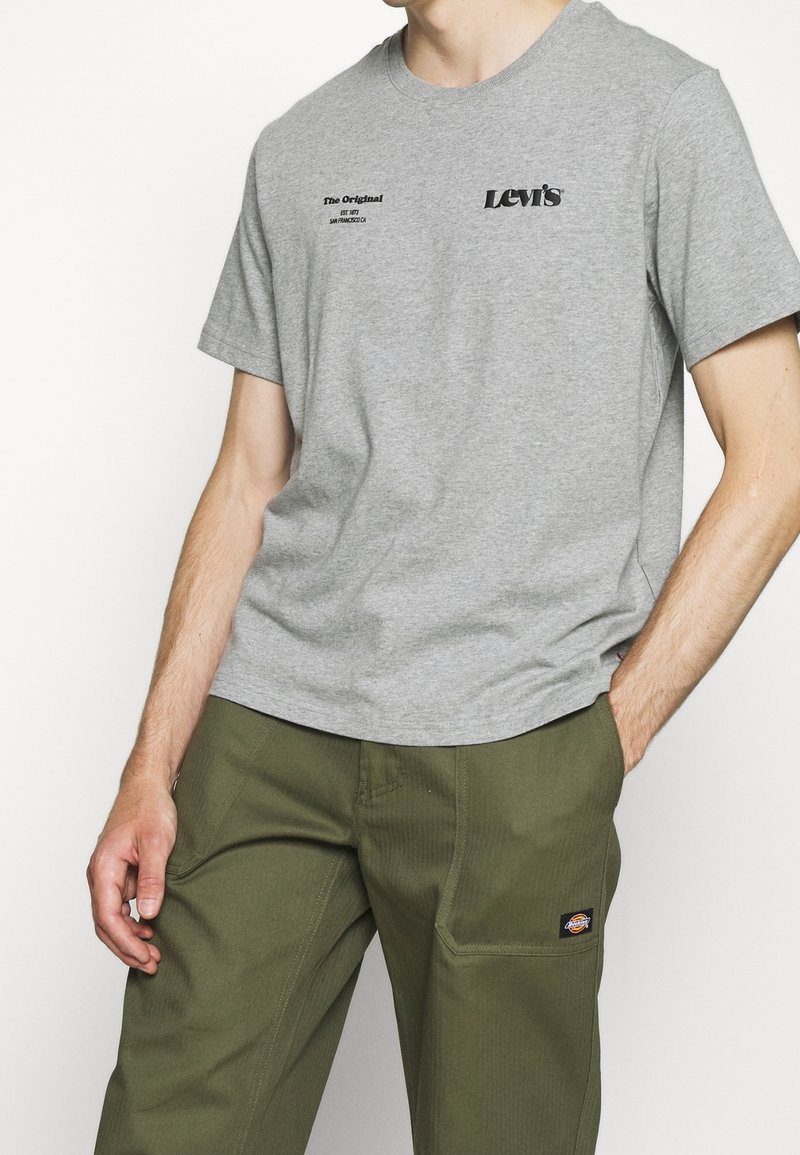 Homme portant un t-shirt Levi's gris avec un texte noir et un pantalon Dickies vert olive avec un patch logo sur la poche, les mains dans les poches.