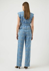 Kobieta z długimi prostymi włosami, ubrana w niebieską dżinsową kamizelkę z haftem i pasujące spodnie, stoi tyłem na białym tle.