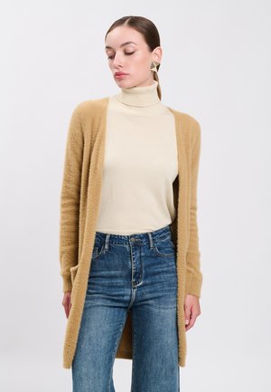 Mujer con suéter de cuello alto beige, cárdigan borroso color camello, vaqueros azules de cintura alta y pendientes geométricos dorados, mirando hacia abajo.