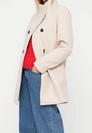 Femme portant un manteau beige à double boutonnage sur un pull rouge et un jean bleu, une main dans la poche, sur un fond clair uni.